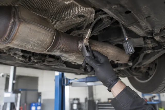 Gloved hand installing an oxygen sensor into an exhaust pipe bung with a sensor socket in a workshop