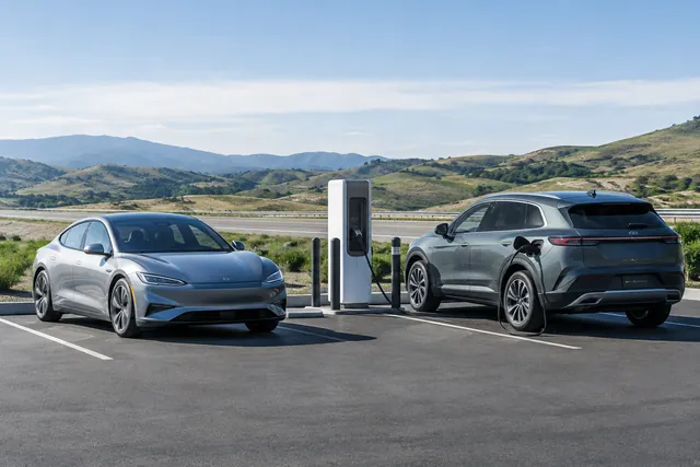 Electric sedan and SUV charging beside a highway rest stop with an open road in the background