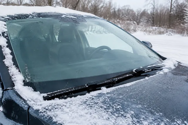 Winter wiper blade resting on a cleared windshield with snow along the edges of a parked car.