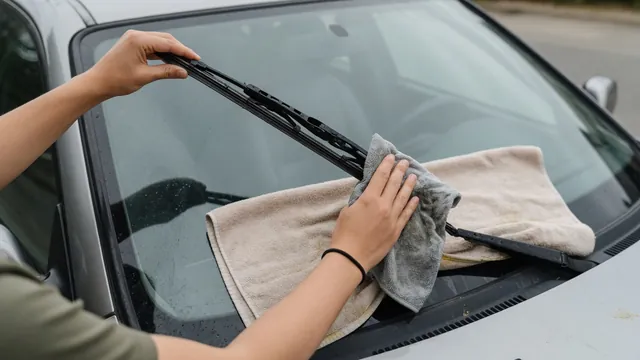 A microfiber cloth cleaning a supported wiper blade beside a parked car’s windshield.