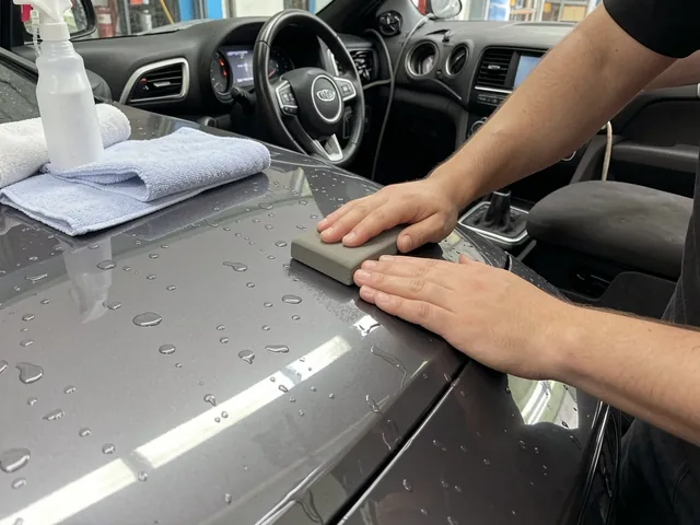 Hands gliding a detailing clay bar over a lubricated car hood beside a clean microfiber towel.