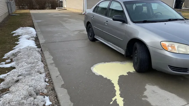 Pale yellow puddle beneath the front of a parked car beside a patch of snow