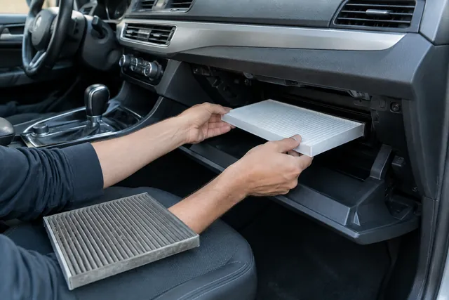Hands installing a new cabin air filter behind a car glove box with the old dirty filter on the seat