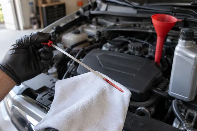 Gloved hand checking the level and color of red automatic transmission fluid on a dipstick with a funnel and fluid bottle nearby