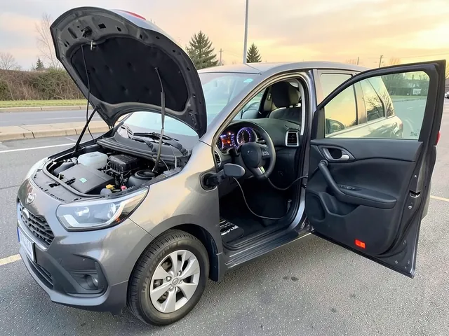Hybrid vehicle with an illuminated dashboard and an open hood showing a covered auxiliary jump terminal.