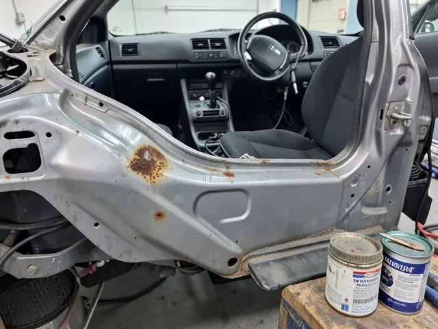 Surface rust on a car body panel beside a brush and an unlabeled rust converter container
