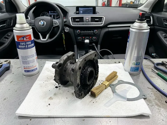 Carbon-coated EGR valve on a workbench next to aerosol cleaner, a brush, a scraper, and a new gasket