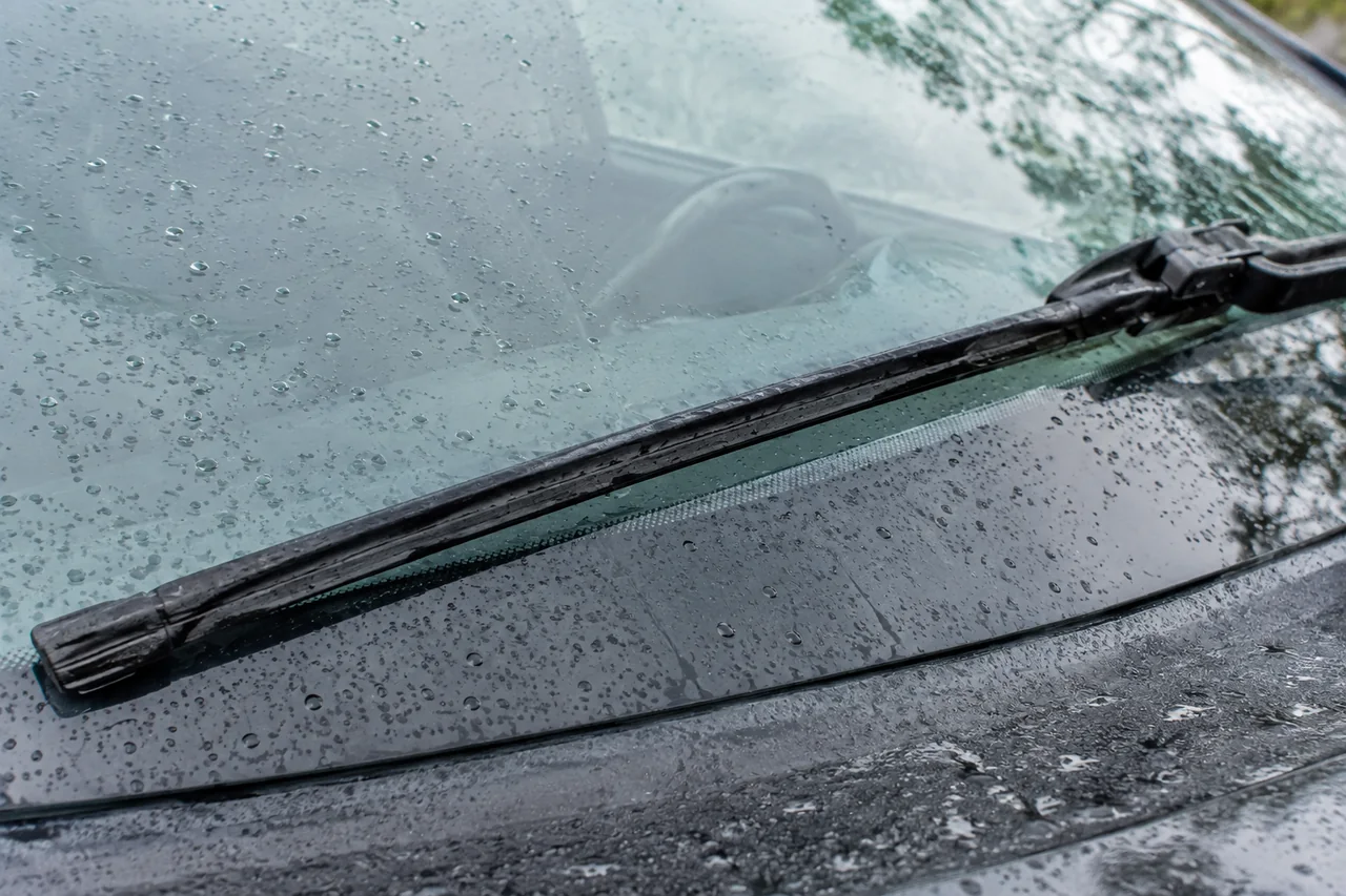Windshield wiper blade with a visibly cracked rubber edge resting on rain-speckled automotive glass.