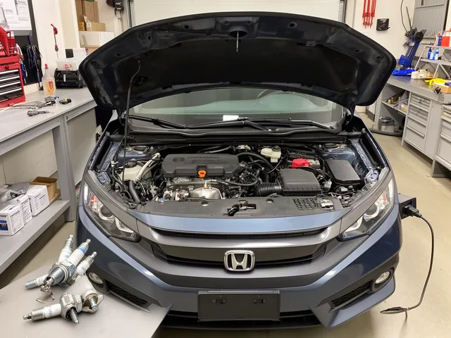 Spark plug and ignition coil arranged on a clean workbench beside an open Honda Civic engine bay