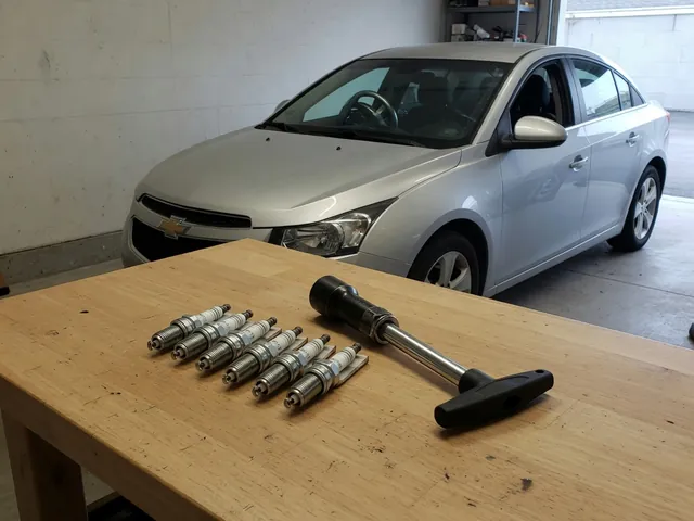 Spark plugs and a spark plug socket arranged on a clean workbench beside a Chevrolet Cruze